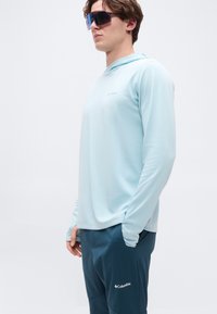 Νέος άνδρας με ανοιχτό μπλε φούτερ με μακριά μανίκια και σκούρο μπλε παντελόνι Columbia, φορώντας μεγάλα αντανακλαστικά αθλητικά γυαλιά ηλίου, ποζάρει μπροστά σε λευκό φόντο.