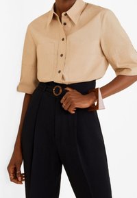 Chemise beige à boutons avec manches courtes, associée à un pantalon noir taille haute et un bracelet manchette rectangulaire marron.