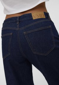 Temno modri denim kavbojke z teksturirano površino. Zasnova vključuje prišite zadnje žepe ter tanek usnjen etiket s "VMD" na pasu.
