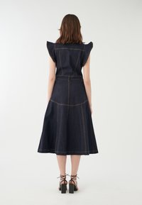 Denimkleid mit kurzen Rüschensäumen, dunkelblauem Stoff, sichtbaren Nähten, ausgestelltem Rock und tailliertem Schnitt. Kombiniert mit schwarzen Blockabsatzsandalen.