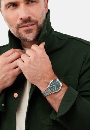 Grüne Baumwolljacke mit hohem Kragen, über einem hellen Hemd getragen. Silberne Metalluhr mit grünem Zifferblatt und silbernem Armband.