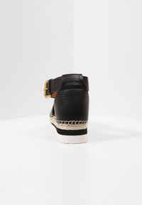 Sandalia de tacón de cuero negro con una hebilla dorada, ribete de yute trenzado y suela blanca, que exhibe una textura suave y un diseño elegante.