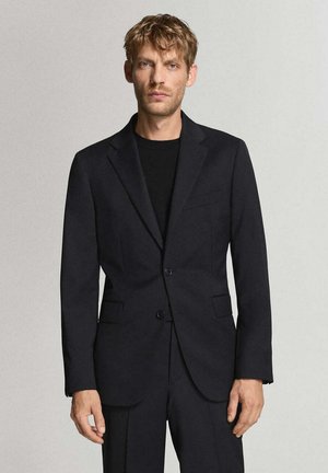 Hombre con cabello castaño claro lleva blazer negro y pantalones a juego sobre camisa negra, de pie frente a un fondo gris liso.