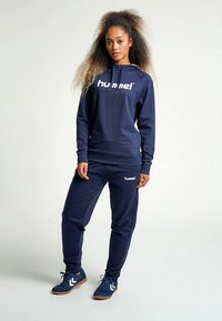 Completo sportivo blu navy composto da una felpa con cappuccio con il logo "hummel", pantaloni abbinati con vita elastica e sneakers blu navy con suola a contrasto.