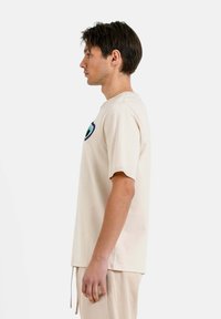 Camiseta de algodón color beige con mangas cortas, que presenta un diseño de corazón bordado en azul y negro en el pecho, con un corte relajado y cuello redondo.