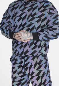 Set di felpa con cappuccio e pantaloni in tessuto iridescente e patterned, caratterizzato da forme angolari nere su uno sfondo blu-verde; include polsini e vita a costine.