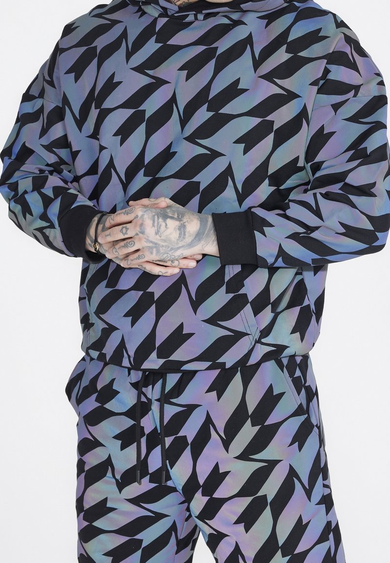 Set di felpa con cappuccio e pantaloni in tessuto iridescente e patterned, caratterizzato da forme angolari nere su uno sfondo blu-verde; include polsini e vita a costine.