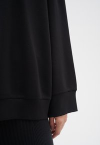 Sudadera negra con una textura suave, presenta un corte holgado, mangas anchas y acentos de canalé en los puños y el dobladillo.