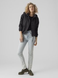 Mlada ženska s kodrastimi blond lasmi, oblečena v črno jakno, črno majico, svetlo modre jeans hlace in temno zelene loaferje, stoji na enobarvnem ozadju.