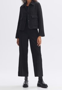 Veste noire texturée avec deux poches avant, fermeture à boutons et col, portée avec un pantalon noir à jambes larges et des bottes épaisses.