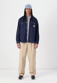 Denim shirt jacket in dark blue, with two chest pockets. Light gray t-shirt underneath. Beige wide-leg pants and dark sneakers. Light blue cap.