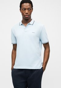 Jeune homme portant un polo bleu clair avec des bordures noires, debout les mains dans les poches de son pantalon bleu marine, regardant sur le côté sur un fond uni.
