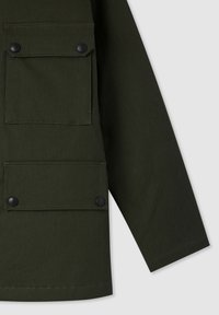 Manica di giacca verde scuro e parte inferiore con due tasche a patta dotate di bottoni a pressione neri su uno sfondo chiaro e uniforme.
