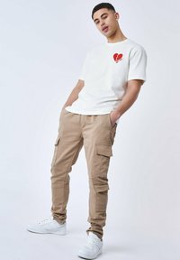 T-shirt bianca con grafica a cuore rosso, abbinata a pantaloni cargo beige con numerose tasche, indossata con sneakers bianche.
