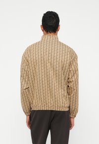 Let let, beige jakke med et geometrisk mønster i mørkebrunt. Har en stående krave, elastiske manchetter og en afslappet pasform.