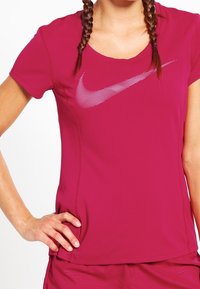 Kvinna med flätat hår iförd en magenta Nike sporttröja och shorts, som vilar en hand på höften mot en vit bakgrund.