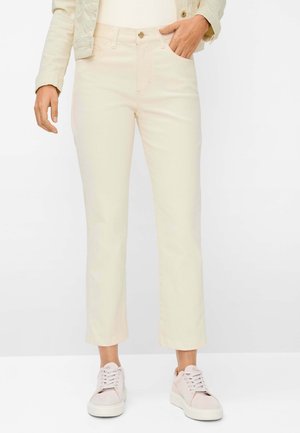 Person iført cremefarvede bukser med lige ben, let jakke og lyserøde sneakers, stående med den ene hånd i lommen mod hvid baggrund.