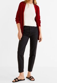 Cardigan en tricot rouge, haut ajusté blanc, jeans noirs à taille haute et sandales plates noires à lanières. La tenue présente une coupe décontractée et confortable.