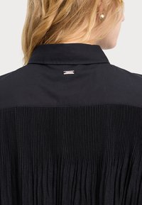 Blusa nera con un design a pieghe sul retro, pannello frontale liscio e un'etichetta argentata con logo al collo.