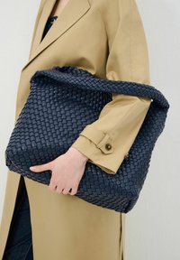 Grote geweven tas van donkerblauw leer. Heeft een gestructureerd, gevlochten ontwerp met een flexibele handgreep. Gestyled met een beige jas en denim.