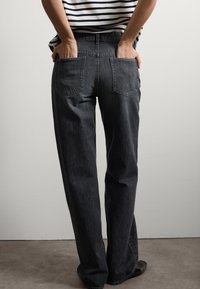 Jeans de mezclilla negra con un diseño de pierna recta. Incluye dos bolsillos traseros y una cintura alta. Llevados con las manos en los bolsillos, con textura mínima.