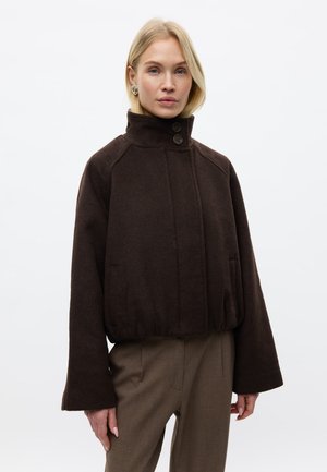 Femme blonde portant une veste en laine marron foncé à col montant avec de gros boutons, des manches larges et un pantalon marron sur mesure, sur un fond uni.