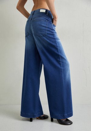 Femme portant un jean bleu taille haute à jambes larges et des chaussures à talons noires, debout avec une main dans la poche sur un fond uni.