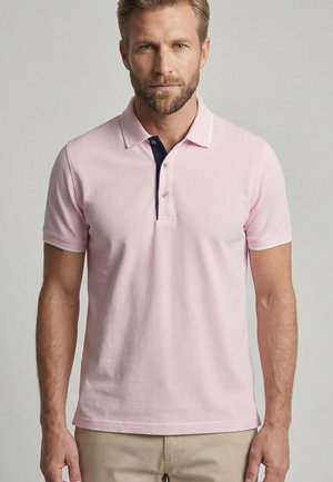 Gebaarde man met een lichtroze poloshirt met korte mouwen, een donkerblauwe binnenbies en beige broek, staand tegen een effen achtergrond.