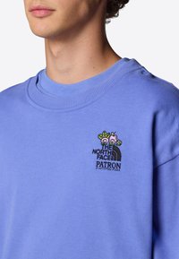 Sudadera azul claro con cuello redondo, que presenta un logotipo bordado en el lado izquierdo del pecho mostrando un diseño floral y el nombre de la marca en negro y verde.
