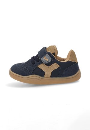 Zapato azul de gamuza con acentos en color tostado, correa de velcro y punta redonda. Suela de goma con superficie texturizada y elementos de diseño en contraste.