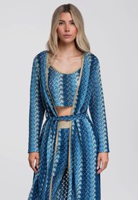 Femme portant un long cardigan bleu à motifs, un crop top assorti et un pantalon avec une bordure dorée métallique sur un fond gris uni.