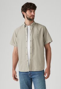 Short-sleeve, light beige button-up shirt, made of cotton, with a pointed collar and straight hem, layered over a white t-shirt.