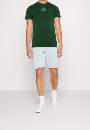 Zielony bawełniany t-shirt z okrągłym dekoltem i krótkimi rękawami, zestawiony z jasnoniebieskimi szortami jeansowymi. Noszony z białymi tenisówkami i widocznymi skarpetkami.