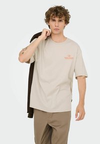 Camiseta de algodón beige con cuello redondo y mangas cortas, que presenta un pequeño logotipo de texto naranja en el pecho. El modelo lleva pantalones marrones y una chaqueta.