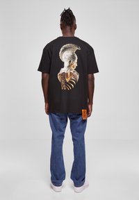 Svart t-skjorte med en detaljert, flerfarget grafikk av en skjelettkriger på ryggen. Rett-sydde jeans og hvite sneakers.