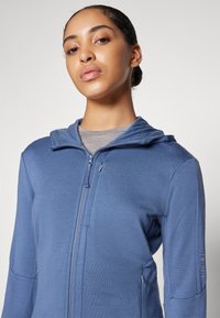 Sweatshirt azul com fecho éclair, feito de tecido texturizado, apresentando um capuz, fecho frontal e bolsos laterais, com um discreto detalhe de logótipo na manga.