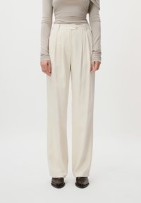 Bredde benbukser i kremfarget stoff, med frontfolder og en glatt tekstur. Stylet med en tettsittende lysebeige langermet topp.