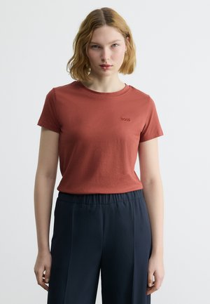 Νεαρή γυναίκα με ξανθό καρέ, φοράει σκουρόχρωμο κόκκινο κοντομάνικο πουκάμισο και σκούρο μπλε παντελόνι ψηλής μέσης, στέκεται μπροστά σε απλό λευκό φόντο.