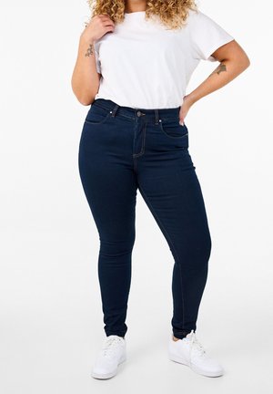 Personne aux cheveux bouclés portant un t-shirt blanc, un jean skinny bleu foncé et des baskets blanches, debout avec une main sur la hanche et l'autre touchant le t-shirt.