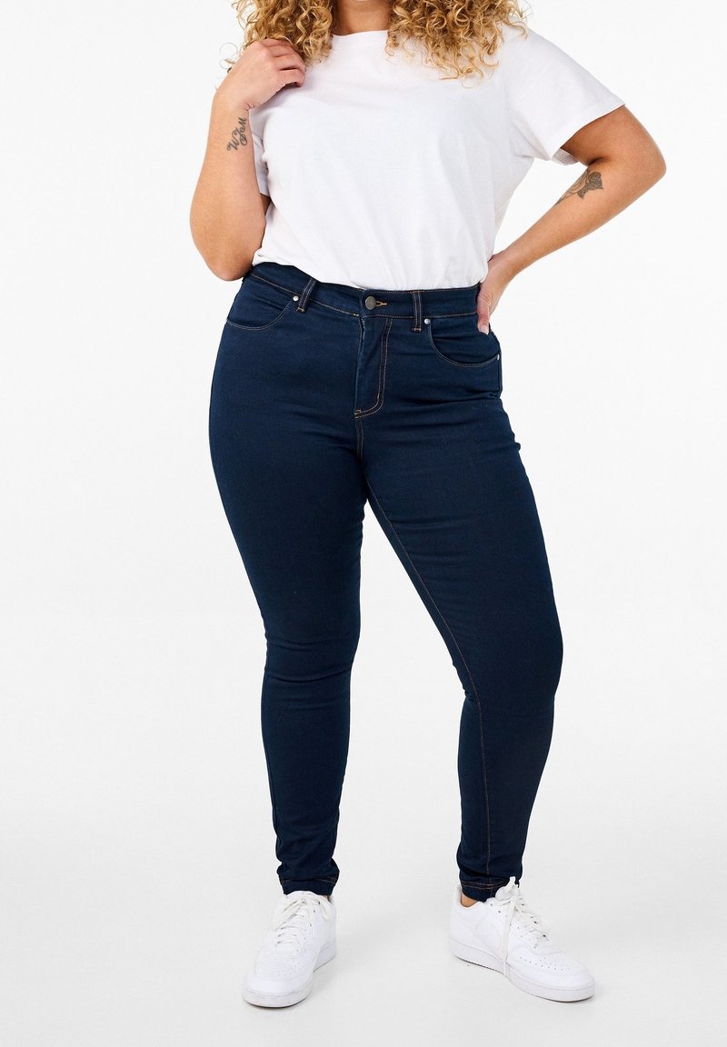 Personne aux cheveux bouclés portant un t-shirt blanc, un jean skinny bleu foncé et des baskets blanches, debout avec une main sur la hanche et l'autre touchant le t-shirt.