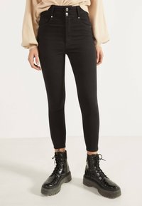 Pantalones ajustados de talle alto en negro, con bolsillos frontales, cierre de botón plateado y ajuste ceñido, combinados con botas de combate negras brillantes con cordones.