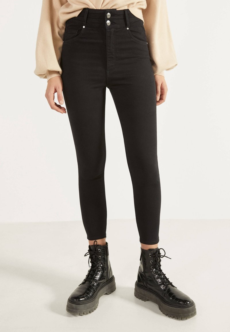 Pantalones ajustados de talle alto en negro, con bolsillos frontales, cierre de botón plateado y ajuste ceñido, combinados con botas de combate negras brillantes con cordones.