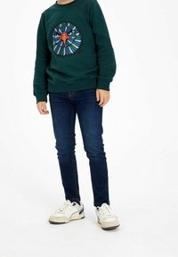 Niño vistiendo un sudadera verde oscura con un diseño colorido de cara de águila, jeans azul oscuro y zapatillas blancas, de pie sobre un fondo blanco.