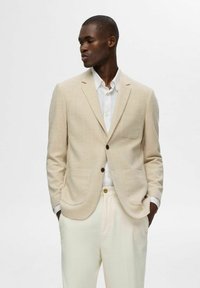 Beżowa marynarka w kratę z jednym kieszonką na piersi, brązowymi guzikami i miękką teksturą; zestawiona z białą koszulą i kremowymi spodniami.