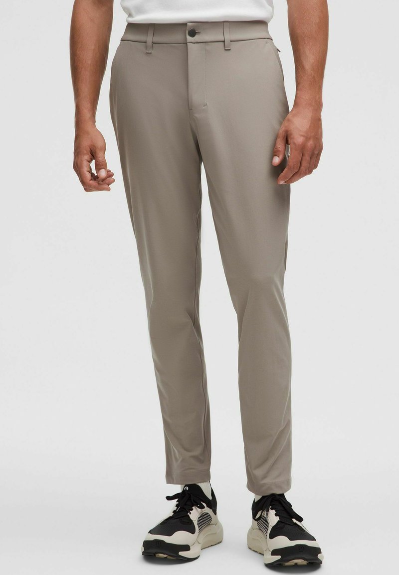 Beżowe, dopasowane spodnie wykonane z gładkiego, elastycznego materiału, z zapięciem na guzik, bocznymi kieszeniami, noszone z czarno-białymi tenisówkami.