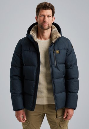 Tmavomodrá prešívaná bunda s kapucňou so kožušinou, so zipsovými vreckami a prešívaným dizajnom. Nosená na béžovom svetri a khaki nohaviciach.