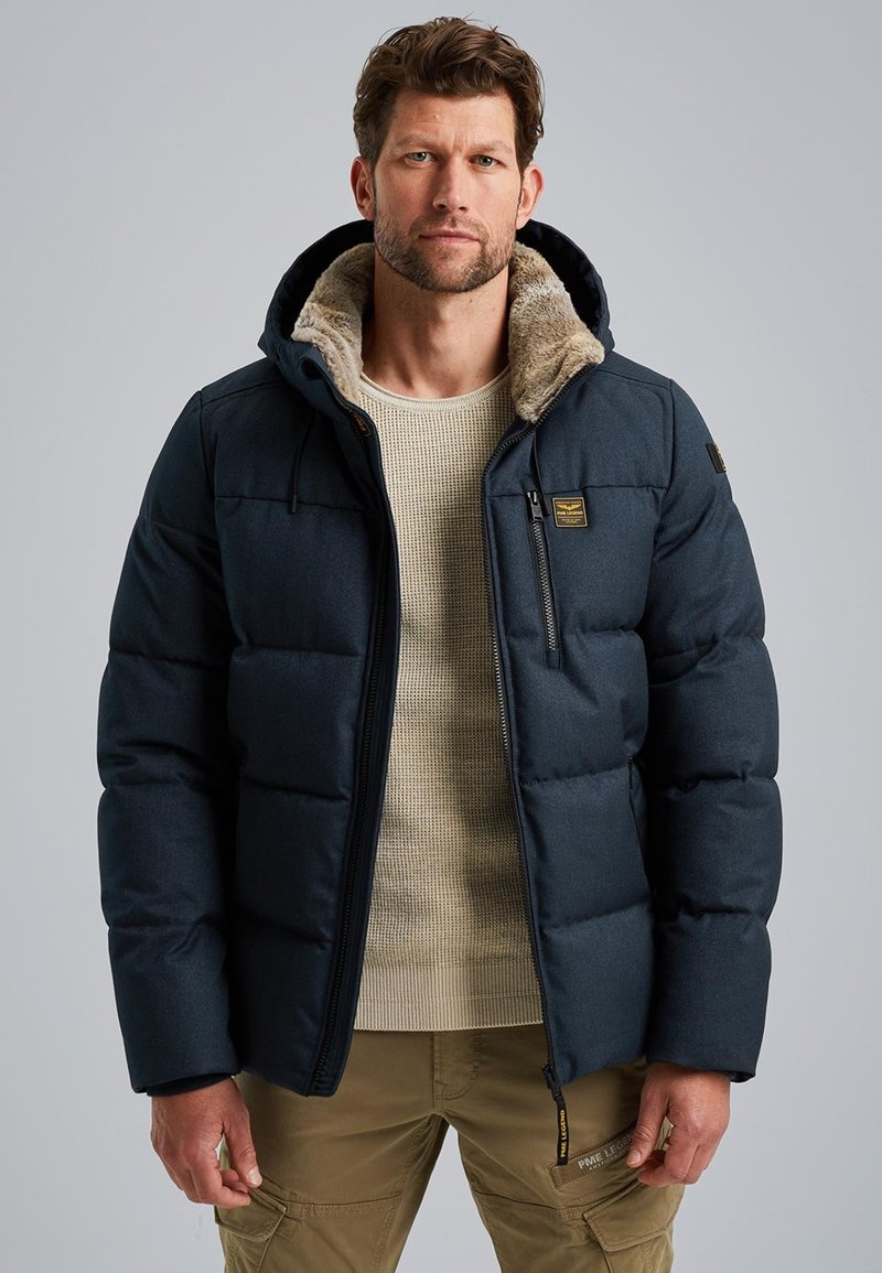 Tmavomodrá prešívaná bunda s kapucňou so kožušinou, so zipsovými vreckami a prešívaným dizajnom. Nosená na béžovom svetri a khaki nohaviciach.