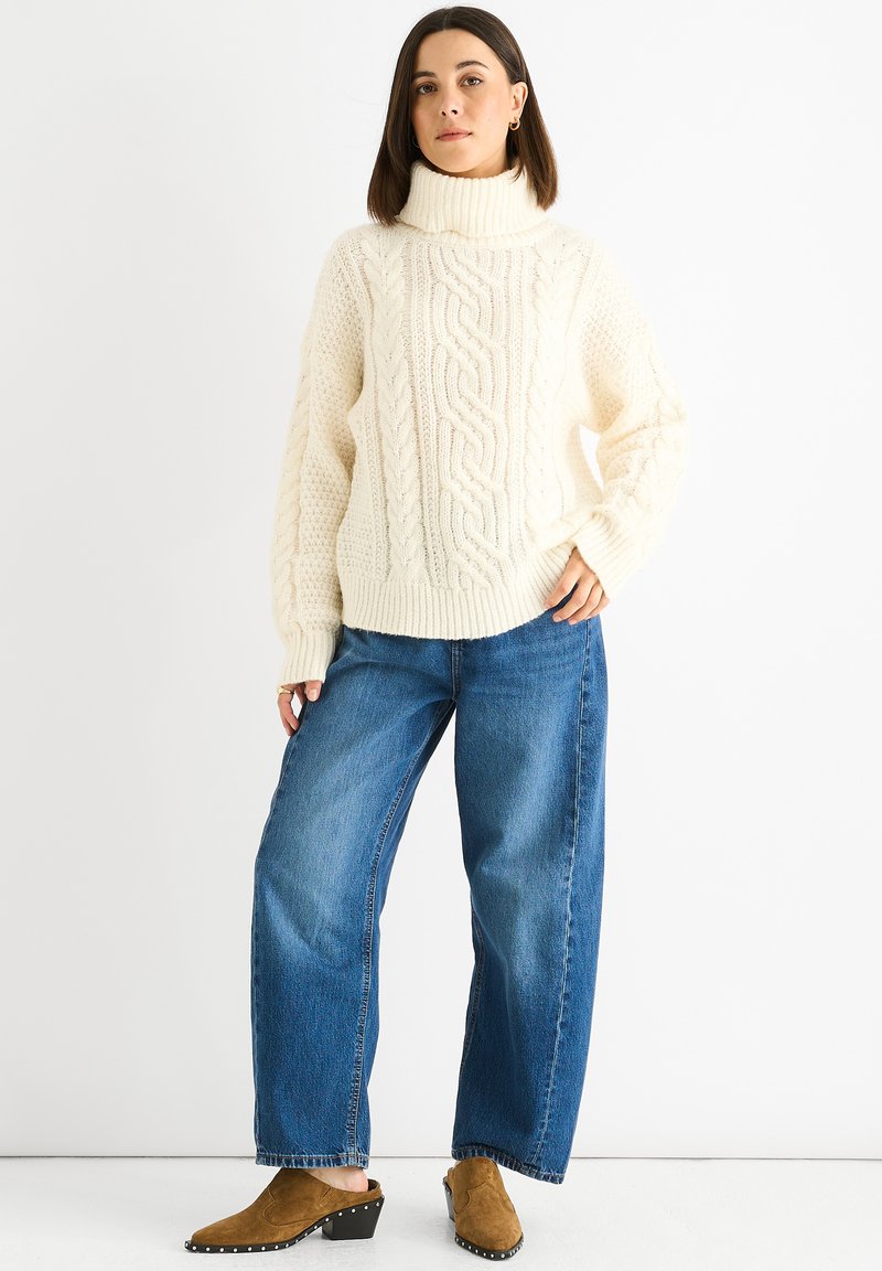 Cream cable-knit turtleneck sweater with chunky texture, paired with high-waisted blue wide-leg jeans and brown heeled mules.