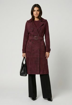 Frau trägt einen langen burgunderroten zweireihigen Trenchcoat mit Gürtel, schwarze weite Hose, schwarze spitze Schuhe, hält eine schwarze Handtasche.