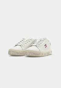 Hvite lær sneakers med en teksturert beige såle, med en fremtredende "TOMMY JEANS" logo og et lite rødt, hvitt og blått emblem på siden.